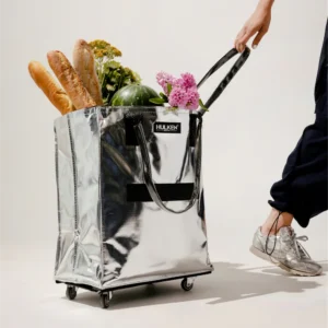 Bags of Brooklyn, Silver Hulken with Groceries poking out the top against a white background while being pulled by a person wearing silver shoes with black pants on.